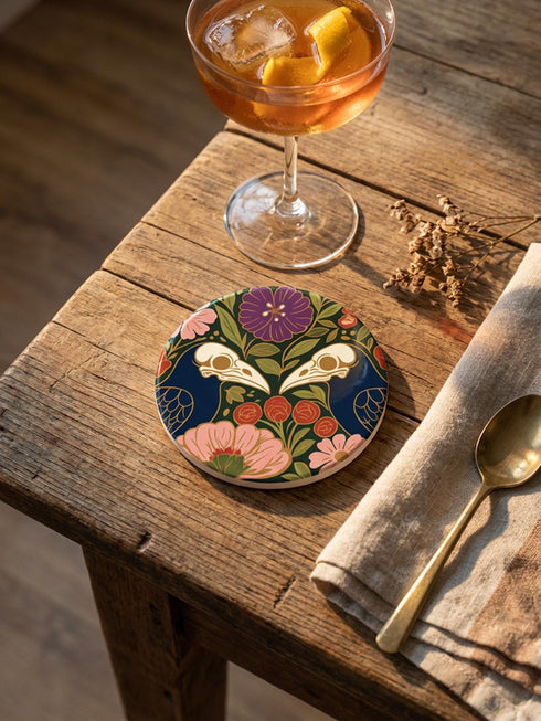 A square drink coaster with a high-gloss finish and natural cork backing. The centralized, symmetrical design features two detailed, cream-colored avian bird skulls facing inward. They are framed by lush botanical flora, including a large purple flower, blush pink blooms, rust-red roses, and green ferns, all set against a deep navy blue background with gold linework accents. Functional gothic, whimsigoth, and dark academia drinkware designed by the Grimoire and Goblet brand.
