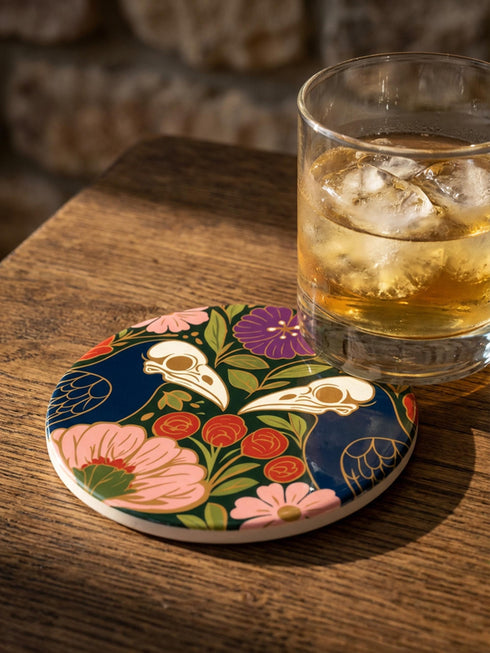 A square drink coaster with a high-gloss finish and natural cork backing. The centralized, symmetrical design features two detailed, cream-colored avian bird skulls facing inward. They are framed by lush botanical flora, including a large purple flower, blush pink blooms, rust-red roses, and green ferns, all set against a deep navy blue background with gold linework accents. Functional gothic, whimsigoth, and dark academia drinkware designed by the Grimoire and Goblet brand.