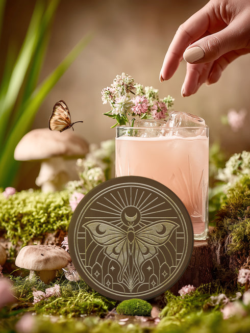A round, dark brown ceramic drink coaster with a non-slip cork back. The top surface features an intricate, engraved-style celestial moth design in a light beige or off-white color. The luna moth is centered under a crescent moon and a radiant sunburst. The design is symmetrical and includes stars and geometric line art, evoking a gothic, witchy, and spiritual aesthetic. The coaster has a matte finish and is designed as a piece of functional art.