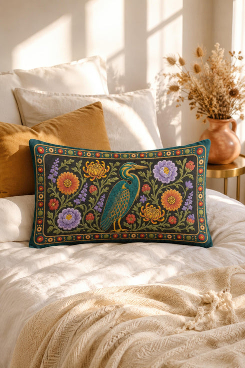 Heron lumbar pillow cover featuring a teal wading bird with gold detailing standing among orange marigolds, golden chrysanthemums, purple peonies, lavender sprigs, and coral red blooms with green foliage on a deep green black panel. A teal border with gold rope trim and a folk style band of red rosettes frames the design. Rectangular 20 x 12 inch cushion case with hidden zipper, the same tapestry look design printed on both sides in smooth, linen feel polyester fabric.