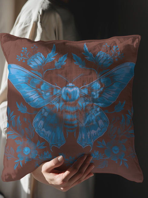 A decorative pillow cover featuring a large Luna moth illustration in electric blue on a rich chocolate brown background, adorned with symmetrical botanical elements and flowers in a detailed vintage naturalist style.