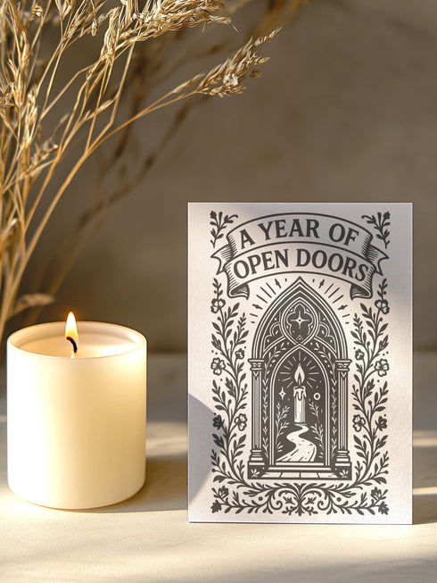 A vertical 5x7 inch pearlescent greeting card by Grimoire and Goblet featuring a high-contrast black woodcut-style illustration. The design centers on a gothic cathedral archway framing a glowing candle and a winding path leading forward. Surrounding the arch are intricate floral vines and stars. A banner at the top reads A YEAR OF OPEN DOORS in serif typography. The paper has a subtle silvery shimmer. The inside contains a printed poem about warmth and promise. Perfect for spiritual gifting.
