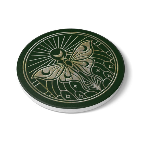 A round, dark hunter green ceramic drink coaster with a non-slip cork back. The top surface features an intricate, engraved-style celestial moth design in a light beige or off-white color. The luna moth is centered under a crescent moon and a radiant sunburst. The design is symmetrical and includes stars and geometric line art, evoking a gothic, witchy, and spiritual aesthetic. The coaster has a matte finish and is designed as a piece of functional art.