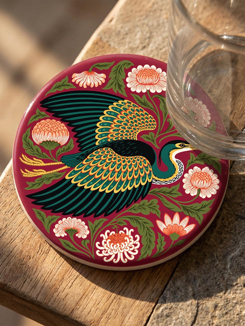 A round, 4-inch ceramic drink coaster featuring a glossy, flat-printed Art Nouveau design. The central motif is a flying heron with teal plumage and scalloped golden wing feathers, surrounded by stylized winding olive green foliage, peach chrysanthemums, and botanical blooms. The artwork is set against a solid, deep blood-red background. The coaster has a smooth white edge and a natural brown cork backing. Available as a single coaster or in matching sets of four or six.
