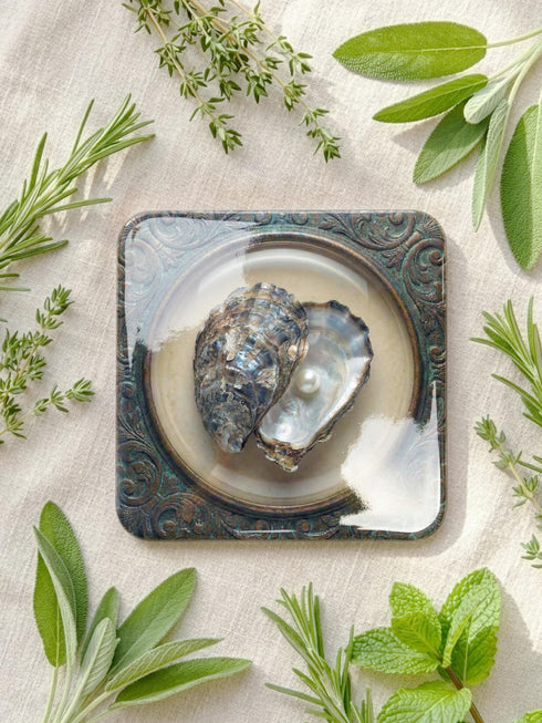 A high-fidelity 2D Trompe-l’œil print on a set of four 4x4 inch square coasters. Each coaster features a centralized sea specimen—a scallop, an oyster with a pearl, a pink conch, and an abalone—centered within an ornate, circular Victorian scrollwork frame. The design uses a faux-texture effect to mimic raised bronze and verdigris metal. The colors include aged gold, teal, and cream, creating a vintage museum specimen aesthetic