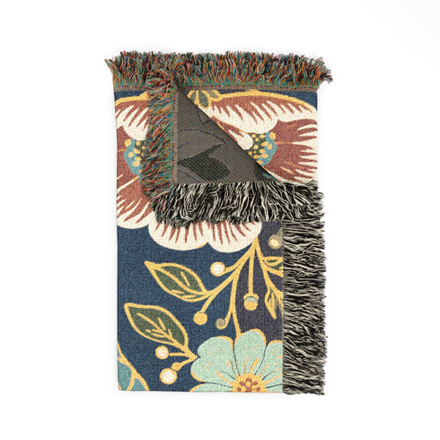 A midnight navy woven cotton blanket featuring a large central raven skull surrounded by rust and ochre flowers, and sage green ferns. Crescent moons are located in the four corners. The blanket has a colorful fringed edge and a textured, loomed appearance. Displayed as a premium gothic botanical home decor piece by Grimoire and Goblet.