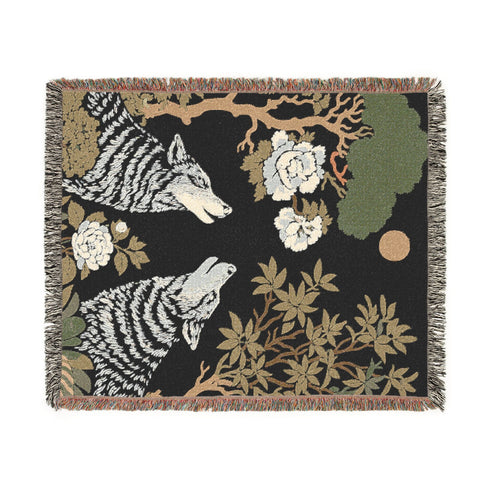 A high-contrast jacquard woven blanket featuring two wolves howling toward a central moon. The design is rendered in charcoal, sage green, and cream threads, showcasing intricate floral motifs like peonies and leafy trees. The textile has a visible woven texture with a multicolored fringe border. This Grimoire and Goblet piece displays a symmetrical, nature-inspired scene that functions as both a heavy throw and a wall tapestry, embodying a dark, botanical aesthetic.