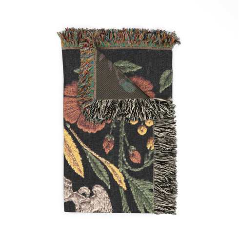 Woven cotton throw blanket with fringed edges and a black ground. Two cream skulls sit side by side at the center of a symmetrical folk garden. A terracotta poppy crowns the top, plum peonies bloom in the middle, and clusters of golden-yellow and white wildflowers, green leaves, and wheat-like sprigs fill the field, with more terracotta poppies along the bottom. The dimensional pattern is woven through a jacquard process, and the reverse shows a mirrored, softer version of the design.