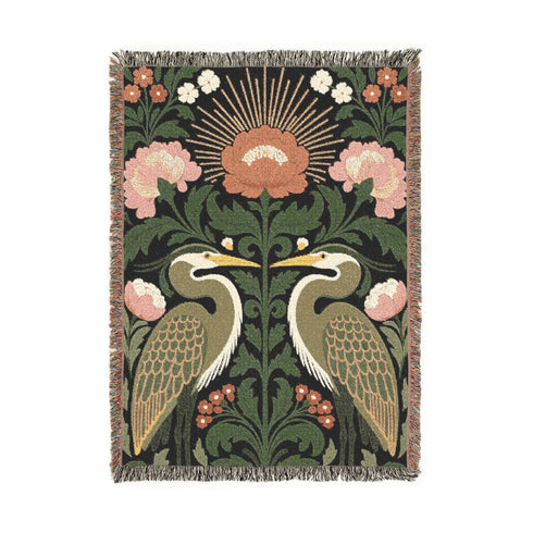 Jacquard woven cotton throw blanket, woven rather than printed, shown flat. Two mirrored great blue herons flank a central radial sunburst, surrounded by botanical blooms and scrollwork. The charcoal black ground carries antique gold ochre sunbeams, blush pink blossoms, deep forest green leaves, muted rust berry clusters, and cream toned herons. A lush multi-colored fringe of black, ochre, and sage threads edges the blanket, and the reverse shows an inverted charcoal grey version of the same design.