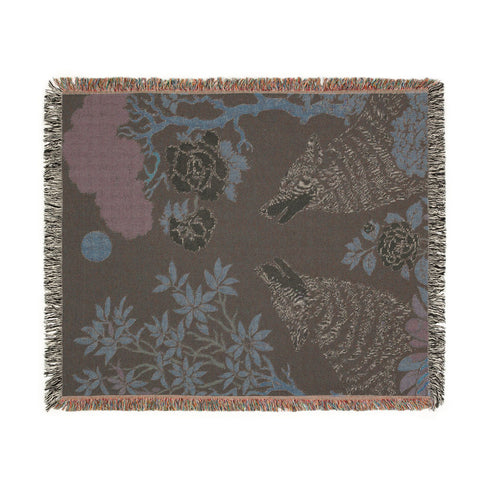 A high-contrast jacquard woven blanket featuring two wolves howling toward a central moon. The design is rendered in charcoal, sage green, and cream threads, showcasing intricate floral motifs like peonies and leafy trees. The textile has a visible woven texture with a multicolored fringe border. This Grimoire and Goblet piece displays a symmetrical, nature-inspired scene that functions as both a heavy throw and a wall tapestry, embodying a dark, botanical aesthetic.
