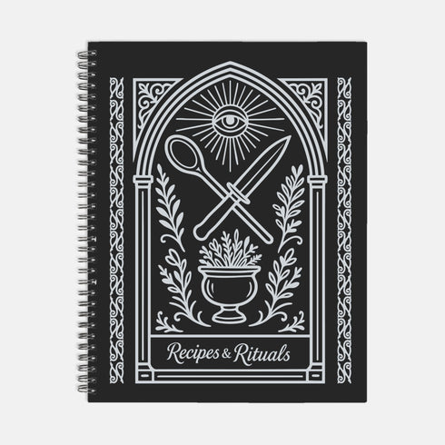 A black spiral-bound hardcover recipe journal lies flat. The cover is matte black with a white line-art design titled "Recipes & Rituals." The art nouveau style illustration features an all-seeing eye under a sunburst, framed by an ornate arch. Below the eye, a crossed spoon and knife are centered above a mortar and pestle filled with herbs. The entire design is bordered by intricate botanical flourishes. The notebook is 8.5 x 11 inches with a wire-o binding, a lay-flat cookbook for kitchen use.