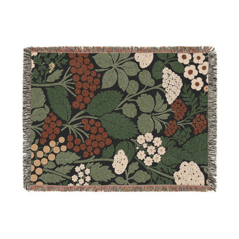 A Grimoire and Goblet luxury woven throw blanket featuring a dark botanical design. The Jacquard cotton tapestry showcases cream elderflower clusters and rust-colored berries against a deep black background. Sage green leaves interweave throughout the floral pattern. The blanket is finished with multicolored fringe edges. A moody cottagecore aesthetic piece ideal for dark academia decor.