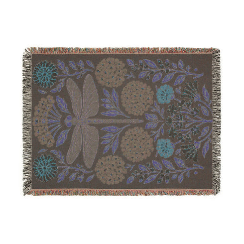 A Grimoire and Goblet Jacquard woven cotton blanket featuring a large central sage green dragonfly. The insect is surrounded by symmetrical clusters of slate blue hydrangeas and muted coral zinnia flowers against a black background. The blanket has a tactile woven texture and is finished with multicolor fringe edges. The design reflects a dark academia and vintage botanical aesthetic.
