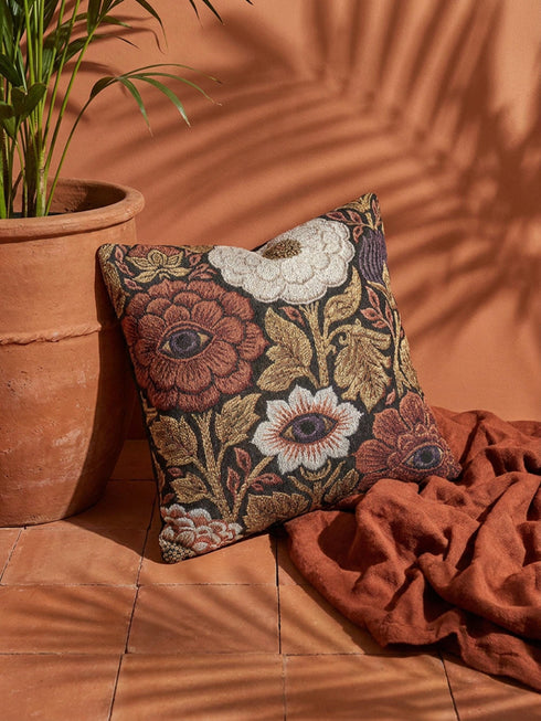 A square woven throw pillow featuring a surreal botanical design on a solid black background. The tapestry-style fabric showcases large, detailed flowers in shades of rust red, cream, and olive green. Nestled within the center of each prominent blossom is a detailed, human-like eye with a dark plum iris, creating a striking evil eye motif. The interwoven threads give the pillow a rich, textured appearance. The pillow is fully stuffed and sewn closed, presenting a smooth, seamless edge.