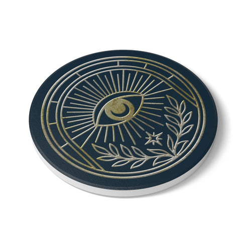 A round ceramic coaster with a smooth, matte, dark slate-blue surface and a natural, textured cork backing. The top of the coaster features an intricate, metallic gold line-art design of the All-Seeing Eye, also known as the Eye of Providence. A central eye with a defined iris and pupil is enclosed within a circular emblem, from which fine golden lines radiate outwards like beams of light. Below the eye, two stylized laurel branches curve upwards, cradling a small, four-pointed star.