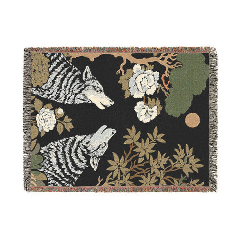 A high-contrast jacquard woven blanket featuring two wolves howling toward a central moon. The design is rendered in charcoal, sage green, and cream threads, showcasing intricate floral motifs like peonies and leafy trees. The textile has a visible woven texture with a multicolored fringe border. This Grimoire and Goblet piece displays a symmetrical, nature-inspired scene that functions as both a heavy throw and a wall tapestry, embodying a dark, botanical aesthetic.