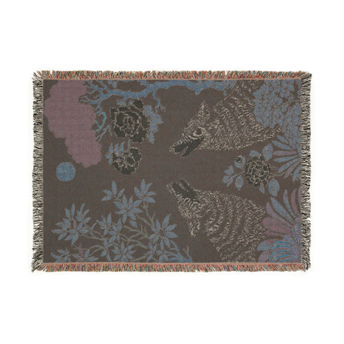 A high-contrast jacquard woven blanket featuring two wolves howling toward a central moon. The design is rendered in charcoal, sage green, and cream threads, showcasing intricate floral motifs like peonies and leafy trees. The textile has a visible woven texture with a multicolored fringe border. This Grimoire and Goblet piece displays a symmetrical, nature-inspired scene that functions as both a heavy throw and a wall tapestry, embodying a dark, botanical aesthetic.