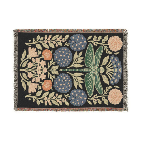 A Grimoire and Goblet Jacquard woven cotton blanket featuring a large central sage green dragonfly. The insect is surrounded by symmetrical clusters of slate blue hydrangeas and muted coral zinnia flowers against a black background. The blanket has a tactile woven texture and is finished with multicolor fringe edges. The design reflects a dark academia and vintage botanical aesthetic.