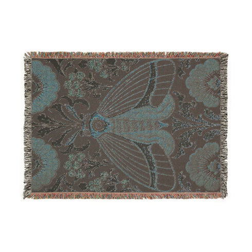 Woven cotton throw blanket with fringed edges and a black ground. At the center is a large moth with copper, brown, and cream wings marked by fine stripes and a concentric eyespot, with feathered antennae. Cream acanthus scrollwork fans out around it in the Art Nouveau manner, joined by clusters of lilac and mauve flowers and copper-brown blossoms with pale rays at the top and bottom. The pattern is jacquard woven, and the reverse shows a mirrored, softer version of the design.