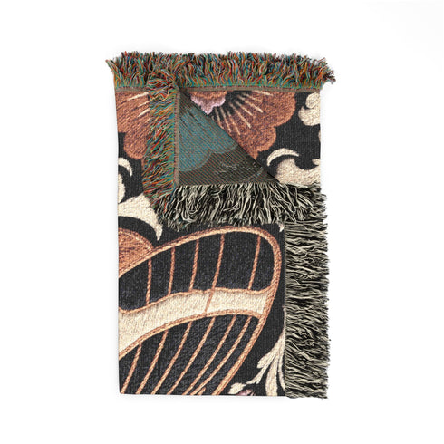 Woven cotton throw blanket with fringed edges and a black ground. At the center is a large moth with copper, brown, and cream wings marked by fine stripes and a concentric eyespot, with feathered antennae. Cream acanthus scrollwork fans out around it in the Art Nouveau manner, joined by clusters of lilac and mauve flowers and copper-brown blossoms with pale rays at the top and bottom. The pattern is jacquard woven, and the reverse shows a mirrored, softer version of the design.