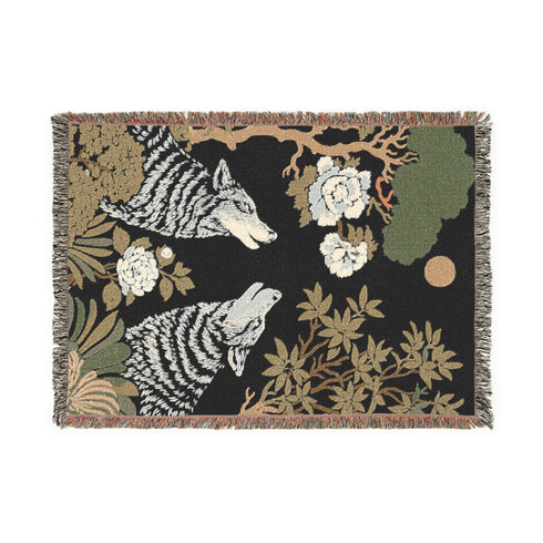 A high-contrast jacquard woven blanket featuring two wolves howling toward a central moon. The design is rendered in charcoal, sage green, and cream threads, showcasing intricate floral motifs like peonies and leafy trees. The textile has a visible woven texture with a multicolored fringe border. This Grimoire and Goblet piece displays a symmetrical, nature-inspired scene that functions as both a heavy throw and a wall tapestry, embodying a dark, botanical aesthetic.