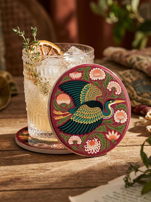 A round, 4-inch ceramic drink coaster featuring a glossy, flat-printed Art Nouveau design. The central motif is a flying heron with teal plumage and scalloped golden wing feathers, surrounded by stylized winding olive green foliage, peach chrysanthemums, and botanical blooms. The artwork is set against a solid, deep blood-red background. The coaster has a smooth white edge and a natural brown cork backing. Available as a single coaster or in matching sets of four or six.