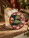 A round, 4-inch ceramic drink coaster featuring a glossy, flat-printed Art Nouveau design. The central motif is a flying heron with teal plumage and scalloped golden wing feathers, surrounded by stylized winding olive green foliage, peach chrysanthemums, and botanical blooms. The artwork is set against a solid, deep blood-red background. The coaster has a smooth white edge and a natural brown cork backing. Available as a single coaster or in matching sets of four or six.