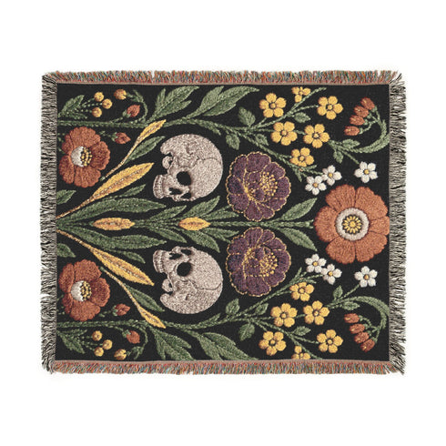 Woven cotton throw blanket with fringed edges and a black ground. Two cream skulls sit side by side at the center of a symmetrical folk garden. A terracotta poppy crowns the top, plum peonies bloom in the middle, and clusters of golden-yellow and white wildflowers, green leaves, and wheat-like sprigs fill the field, with more terracotta poppies along the bottom. The dimensional pattern is woven through a jacquard process, and the reverse shows a mirrored, softer version of the design.