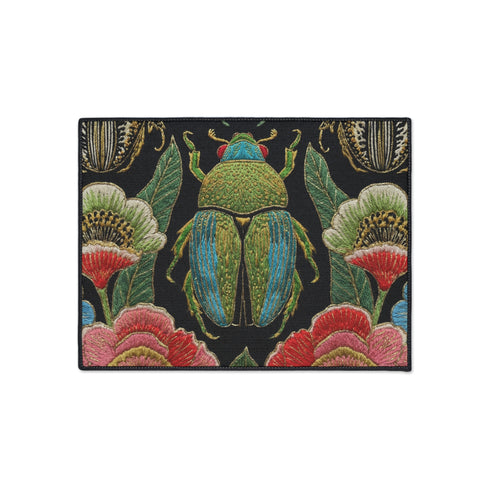 A rectangular doormat featuring a high-fidelity faux embroidery print on a solid black background. The central design is a large, detailed green and teal scarab beetle. It is surrounded by vibrant, flat-printed botanical motifs including large pink, red, and light blue flowers, green foliage, and pale gold-toned seed pods. The mat features a black fabric trim around the edges. The colorful print creates a 2D trompe-l'œil illusion of raised tapestry needlework on a smooth polyester surface.