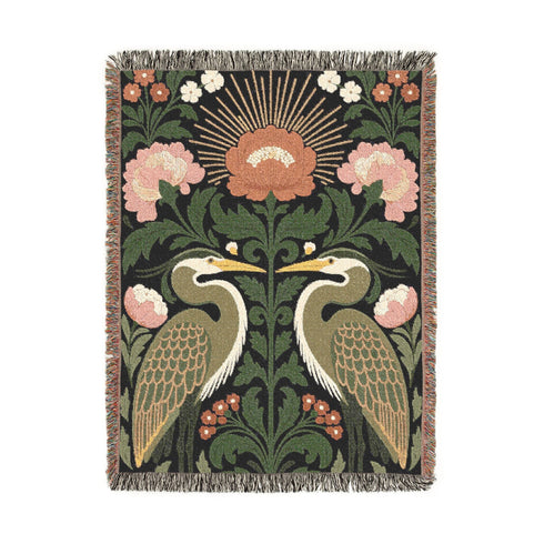 Jacquard woven cotton throw blanket, woven rather than printed, shown flat. Two mirrored great blue herons flank a central radial sunburst, surrounded by botanical blooms and scrollwork. The charcoal black ground carries antique gold ochre sunbeams, blush pink blossoms, deep forest green leaves, muted rust berry clusters, and cream toned herons. A lush multi-colored fringe of black, ochre, and sage threads edges the blanket, and the reverse shows an inverted charcoal grey version of the same design.