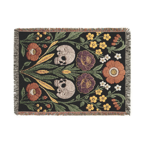 Woven cotton throw blanket with fringed edges and a black ground. Two cream skulls sit side by side at the center of a symmetrical folk garden. A terracotta poppy crowns the top, plum peonies bloom in the middle, and clusters of golden-yellow and white wildflowers, green leaves, and wheat-like sprigs fill the field, with more terracotta poppies along the bottom. The dimensional pattern is woven through a jacquard process, and the reverse shows a mirrored, softer version of the design.