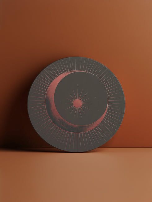 A round ceramic plate with a sleek design featuring a crescent moon and radiating lines in a muted red and black color scheme. The plate is set against a warm brown background with soft lighting, highlighting the celestial and minimalist aesthetic of the piece.