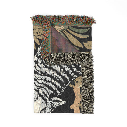 A high-contrast jacquard woven blanket featuring two wolves howling toward a central moon. The design is rendered in charcoal, sage green, and cream threads, showcasing intricate floral motifs like peonies and leafy trees. The textile has a visible woven texture with a multicolored fringe border. This Grimoire and Goblet piece displays a symmetrical, nature-inspired scene that functions as both a heavy throw and a wall tapestry, embodying a dark, botanical aesthetic.
