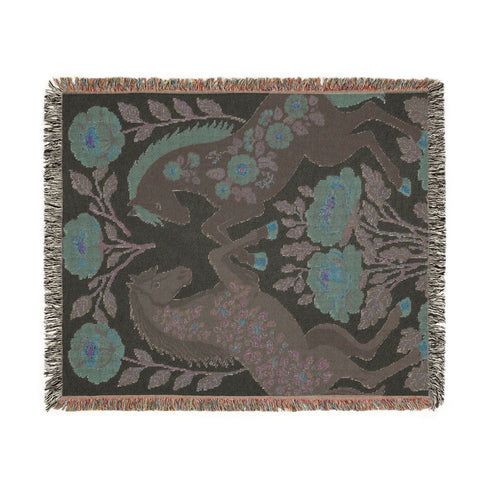 A woven cotton throw blanket by Grimoire and Goblet featuring two rearing horses facing each other in a symmetrical folk art design. One horse is charcoal black with rust floral detailing; the other is navy blue with teal floral detailing. They are framed by large rust-colored peonies and green leafy vines on a cream background. The blanket features a multicolor fringe edge. Perfect for dark academia and cottagecore aesthetics.