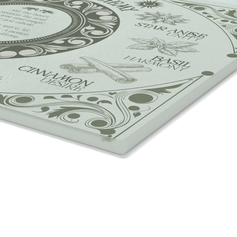 A rectangular sage-tinted glass cutting board featuring an ornate design of magical herbs and spices. Text reads "Love's Alchemy" with detailed illustrations of vanilla, rose, cardamom, star anise, basil, and cinnamon arranged around a central medallion containing a love spell verse.