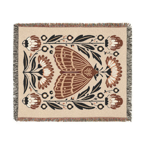 Grimoire and Goblet woven jacquard cotton blanket / wall tapestry featuring a centered folk-art moth framed by symmetrical wildflowers and leafy stems. Natural oatmeal-beige base with rust/terracotta and cocoa-brown moth detail, charcoal-black botanicals, and warm ivory highlights. Finished with a 2-inch multicolor fringe and a reversible color-negative back. Available in 52x37, 50x60, and 60x80 inches as a throw, bed cover, or statement wall hanging.