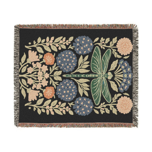A Grimoire and Goblet Jacquard woven cotton blanket featuring a large central sage green dragonfly. The insect is surrounded by symmetrical clusters of slate blue hydrangeas and muted coral zinnia flowers against a black background. The blanket has a tactile woven texture and is finished with multicolor fringe edges. The design reflects a dark academia and vintage botanical aesthetic.