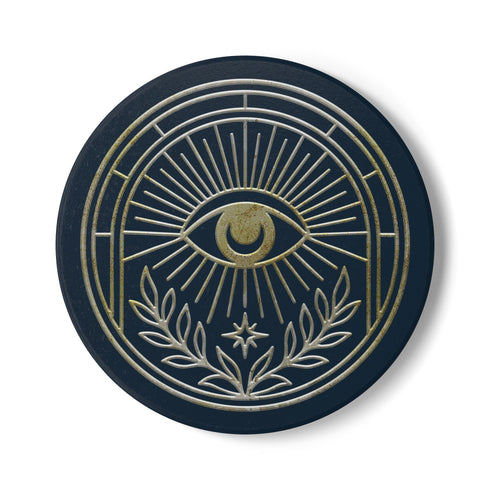 A round ceramic coaster with a smooth, matte, dark slate-blue surface and a natural, textured cork backing. The top of the coaster features an intricate, metallic gold line-art design of the All-Seeing Eye, also known as the Eye of Providence. A central eye with a defined iris and pupil is enclosed within a circular emblem, from which fine golden lines radiate outwards like beams of light. Below the eye, two stylized laurel branches curve upwards, cradling a small, four-pointed star.