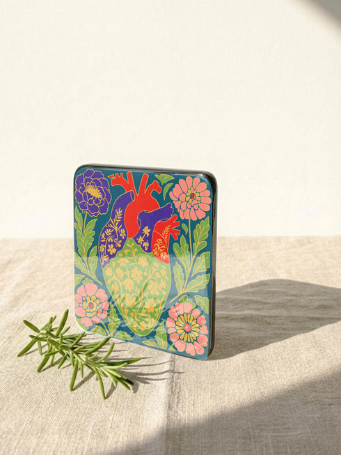A set of four premium 4x4 inch anatomical heart coasters from Grimoire and Goblet. Each square coaster is made of masonite hardboard with a high-gloss finish and a non-slip cork base. The designs feature intricate biological heart illustrations with folk-art floral motifs in vibrant crimson, pink, yellow, and purple against mossy green and deep teal backgrounds. The high-fidelity print mimics a luxury embroidered texture, perfect for a cardiology gift or dark academia home decor.