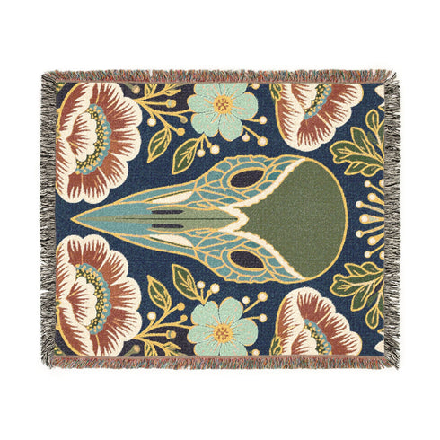 A midnight navy woven cotton blanket featuring a large central raven skull surrounded by rust and ochre flowers, and sage green ferns. Crescent moons are located in the four corners. The blanket has a colorful fringed edge and a textured, loomed appearance. Displayed as a premium gothic botanical home decor piece by Grimoire and Goblet.