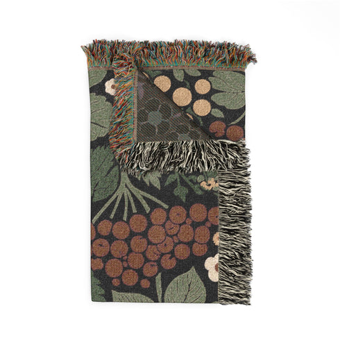 A Grimoire and Goblet luxury woven throw blanket featuring a dark botanical design. The Jacquard cotton tapestry showcases cream elderflower clusters and rust-colored berries against a deep black background. Sage green leaves interweave throughout the floral pattern. The blanket is finished with multicolored fringe edges. A moody cottagecore aesthetic piece ideal for dark academia decor.