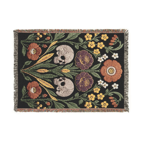Woven cotton throw blanket with fringed edges and a black ground. Two cream skulls sit side by side at the center of a symmetrical folk garden. A terracotta poppy crowns the top, plum peonies bloom in the middle, and clusters of golden-yellow and white wildflowers, green leaves, and wheat-like sprigs fill the field, with more terracotta poppies along the bottom. The dimensional pattern is woven through a jacquard process, and the reverse shows a mirrored, softer version of the design.