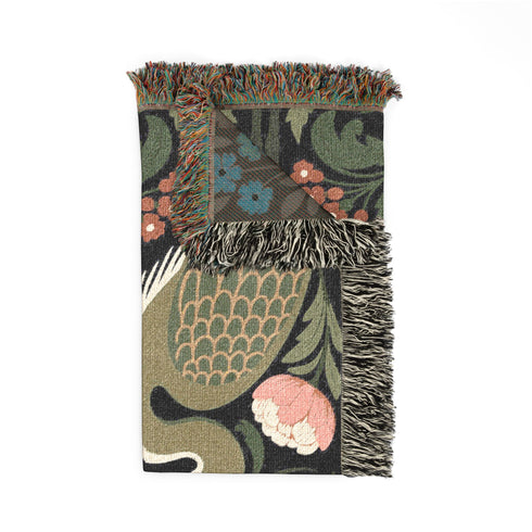 Jacquard woven cotton throw blanket, woven rather than printed, shown flat. Two mirrored great blue herons flank a central radial sunburst, surrounded by botanical blooms and scrollwork. The charcoal black ground carries antique gold ochre sunbeams, blush pink blossoms, deep forest green leaves, muted rust berry clusters, and cream toned herons. A lush multi-colored fringe of black, ochre, and sage threads edges the blanket, and the reverse shows an inverted charcoal grey version of the same design.