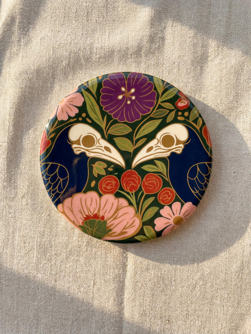 A square drink coaster with a high-gloss finish and natural cork backing. The centralized, symmetrical design features two detailed, cream-colored avian bird skulls facing inward. They are framed by lush botanical flora, including a large purple flower, blush pink blooms, rust-red roses, and green ferns, all set against a deep navy blue background with gold linework accents. Functional gothic, whimsigoth, and dark academia drinkware designed by the Grimoire and Goblet brand.