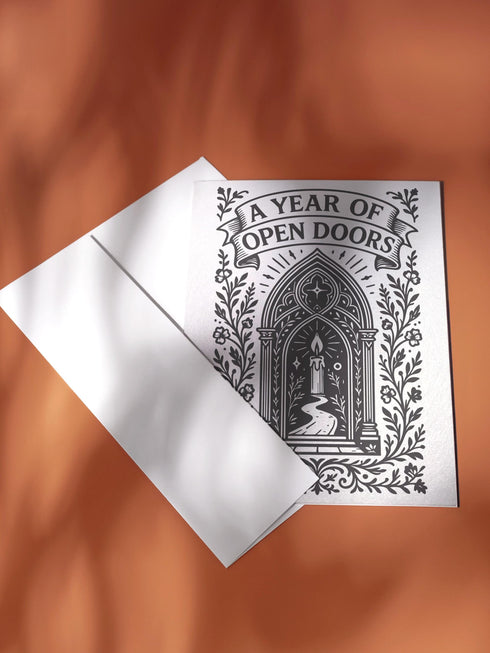 A vertical 5x7 inch pearlescent greeting card by Grimoire and Goblet featuring a high-contrast black woodcut-style illustration. The design centers on a gothic cathedral archway framing a glowing candle and a winding path leading forward. Surrounding the arch are intricate floral vines and stars. A banner at the top reads A YEAR OF OPEN DOORS in serif typography. The paper has a subtle silvery shimmer. The inside contains a printed poem about warmth and promise. Perfect for spiritual gifting.