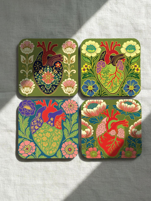 A set of four premium 4x4 inch anatomical heart coasters from Grimoire and Goblet. Each square coaster is made of masonite hardboard with a high-gloss finish and a non-slip cork base. The designs feature intricate biological heart illustrations with folk-art floral motifs in vibrant crimson, pink, yellow, and purple against mossy green and deep teal backgrounds. The high-fidelity print mimics a luxury embroidered texture, perfect for a cardiology gift or dark academia home decor.