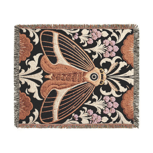 Woven cotton throw blanket with fringed edges and a black ground. At the center is a large moth with copper, brown, and cream wings marked by fine stripes and a concentric eyespot, with feathered antennae. Cream acanthus scrollwork fans out around it in the Art Nouveau manner, joined by clusters of lilac and mauve flowers and copper-brown blossoms with pale rays at the top and bottom. The pattern is jacquard woven, and the reverse shows a mirrored, softer version of the design.