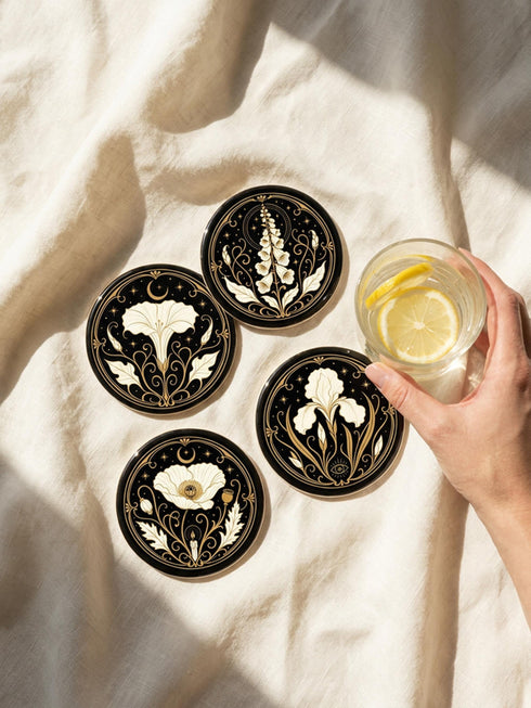 A set of four round 4-inch ceramic coasters featuring a glossy 2D flat-printed Art Deco botanical design. Each coaster displays a different cream-colored flower including a moonflower, foxglove, poppy, and iris, set against a solid black background. The floral motifs are framed by intricate gold linework, crescent moons, four-pointed stars, and mystical symbols like a candle and an all-seeing eye. The coasters have a smooth white edge and are finished with a natural brown cork backing.