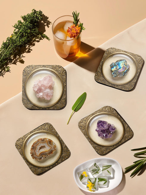 Grimoire and Goblet set of four square glossy coasters featuring high-resolution printed illustrations of raw crystals: purple amethyst, pink rose quartz, iridescent labradorite, and banded agate. Each gemstone is depicted inside a printed faux antique brass baroque frame with intricate scrollwork, set against a cream parchment background. The design is a flat 2D trompe l'œil print on hardboard, creating a realistic 3D specimen box illusion.