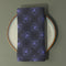 Set of four charcoal-colored cloth napkins with a repeating pattern of triangular all-seeing eyes in purple, surrounded by radiating lines and small star-like accents.